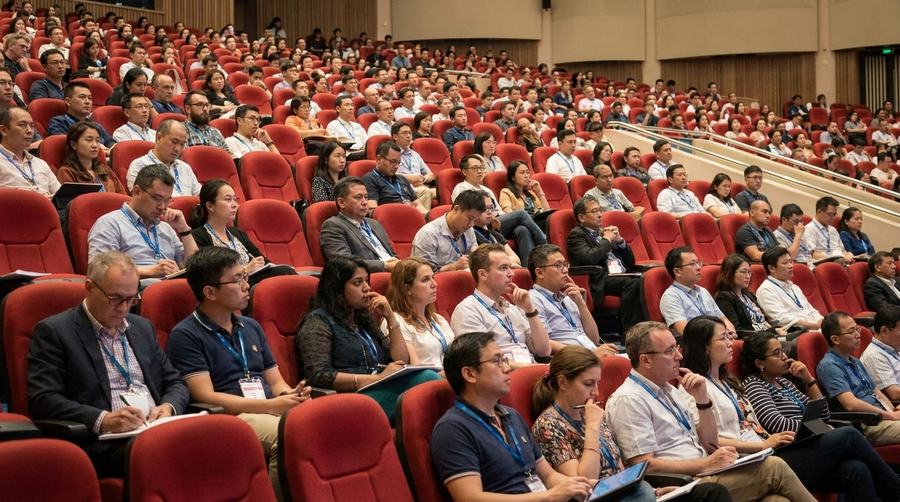A conference audience seated in red chairs, taking notes