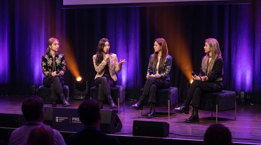 Four speakers on a panel stage with an audience in front
